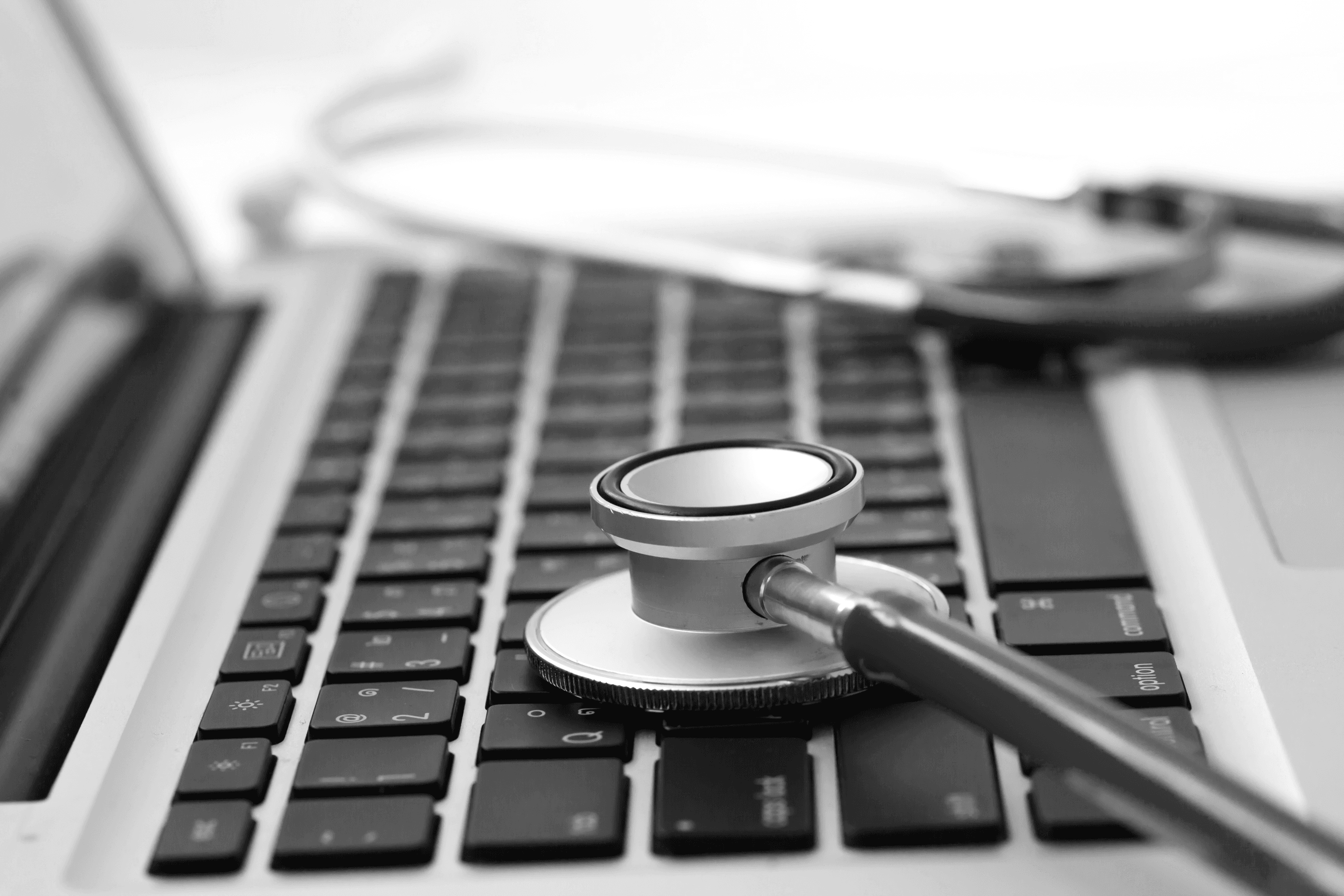 A stethoscope rests on the keyboard of a laptop. The instrument is silver with a blue tube, and a small portion of the white background can be seen in the background.